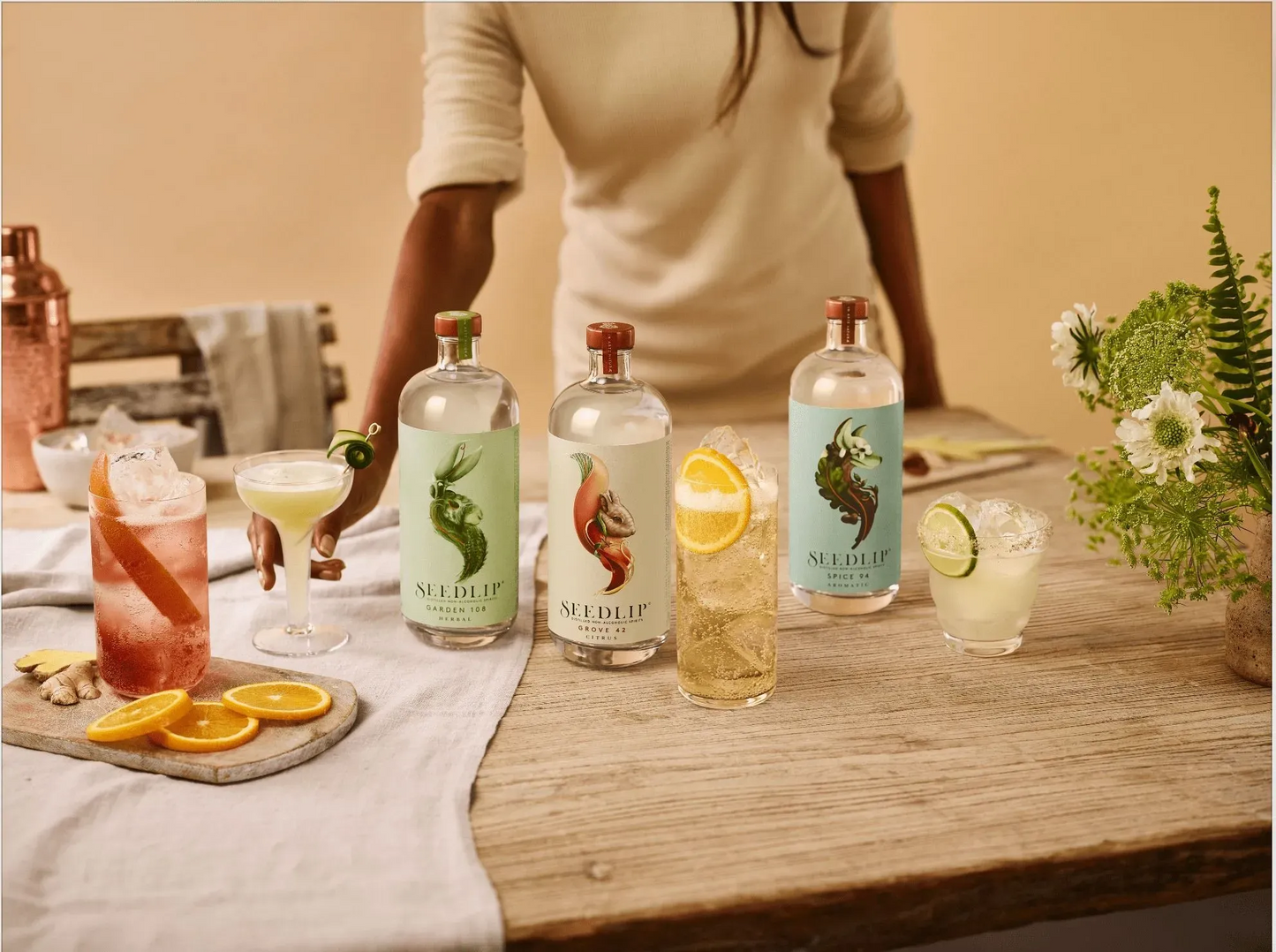 Table with four bottles, cocktails, and garnishes with a person partially visible.