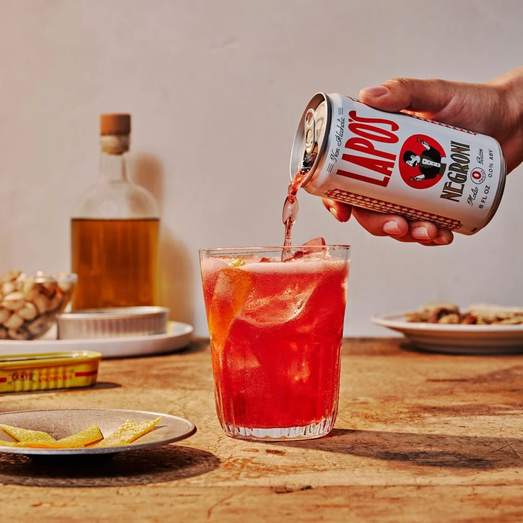Person pouring a red cocktail from a can labeled 'Lapos' into a glass on a wooden table.