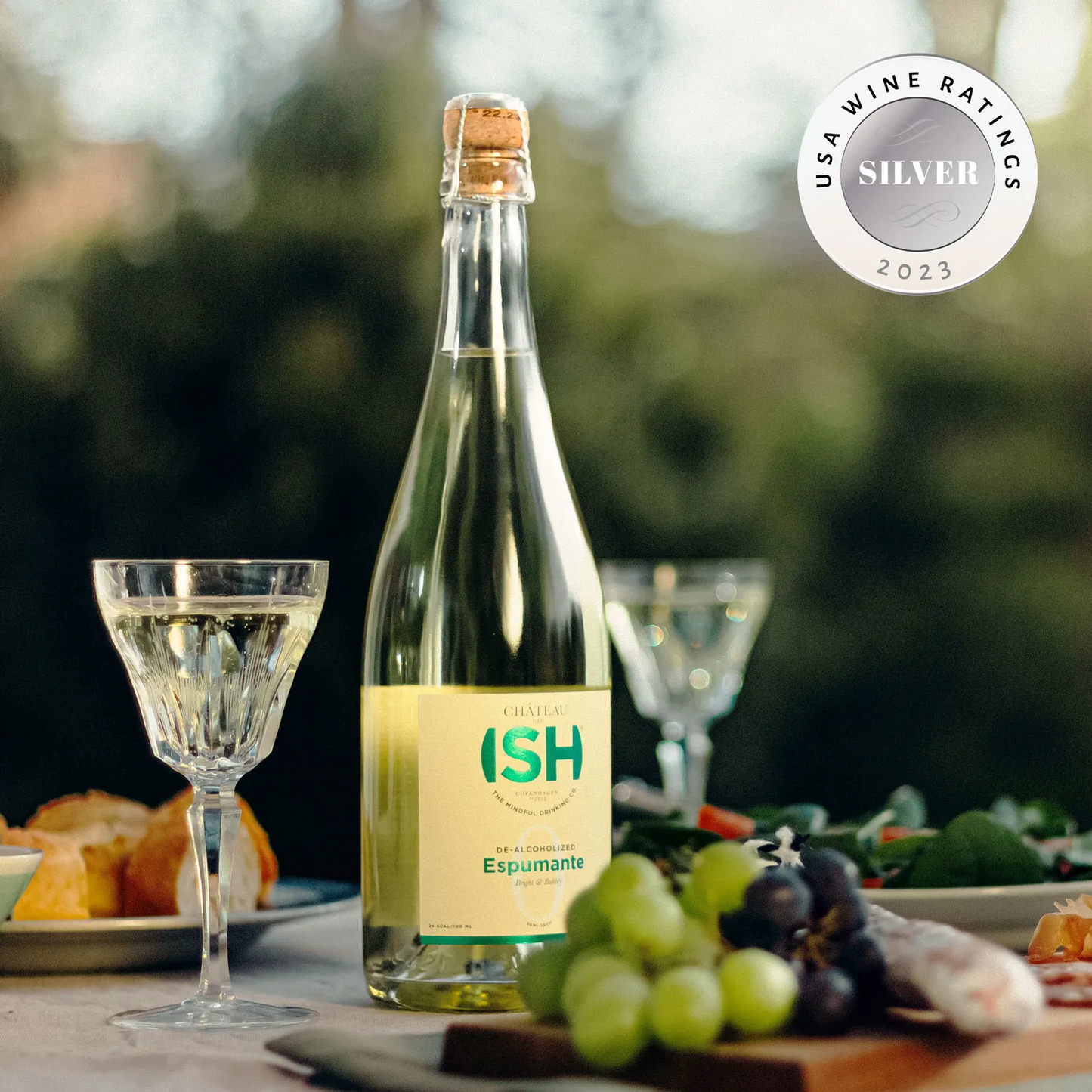 Bottle of Ish Espumante with a glass of wine on a table outdoors, featuring a silver USA Wine Ratings 2023 badge.