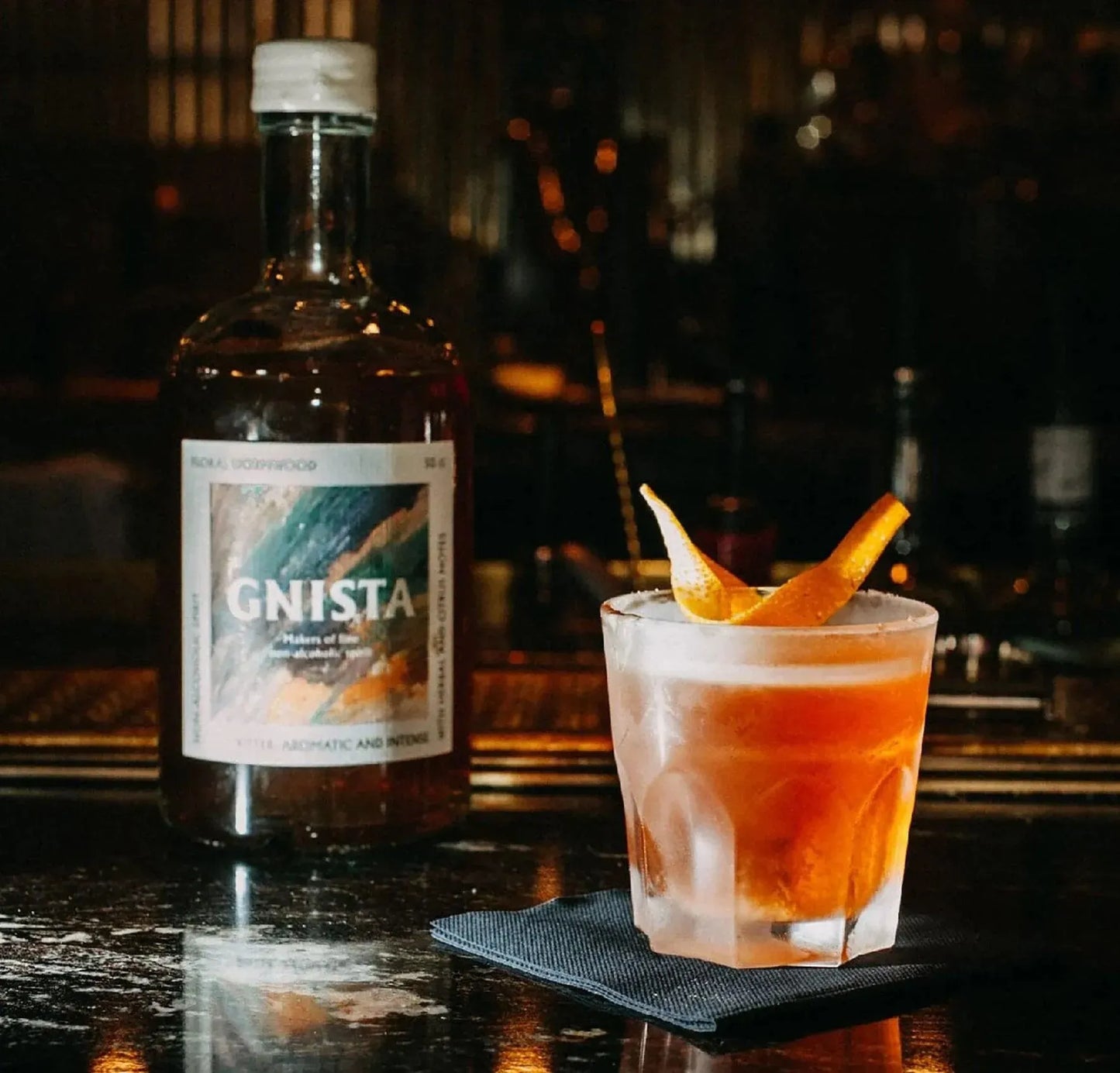 Bottle of GNISTA whiskey and a cocktail with an orange twist on a bar.