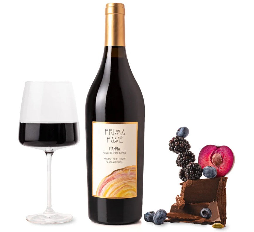A bottle of Prima Pavé red wine stands next to a filled wine glass, with chocolate, berries, and a sliced plum arranged beside it on a clean white background.