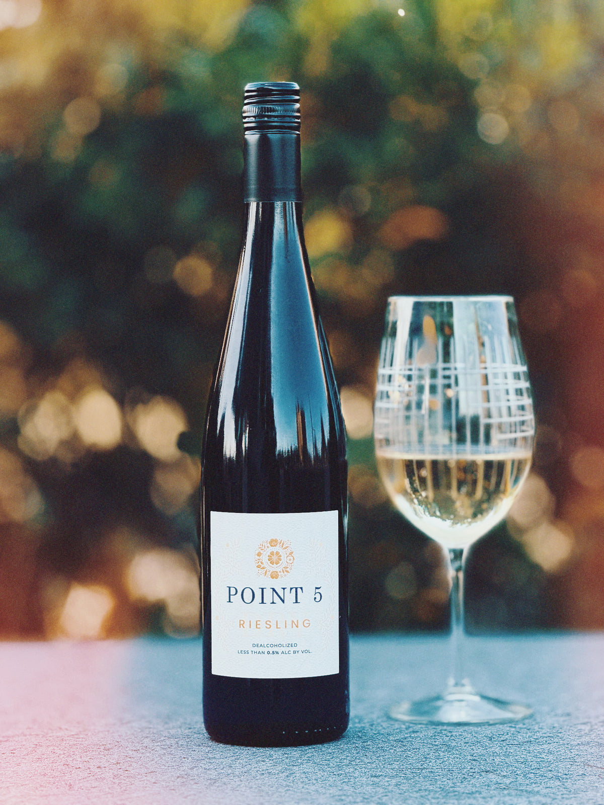 A bottle of Point 5 Riesling wine stands on a stone surface next to a filled wine glass, with a softly blurred outdoor garden background.