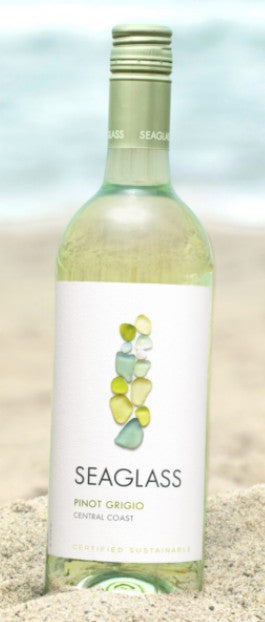 A bottle of Seaglass Pinot Grigio Central Coast white wine standing upright in sand with a blurred beach background.