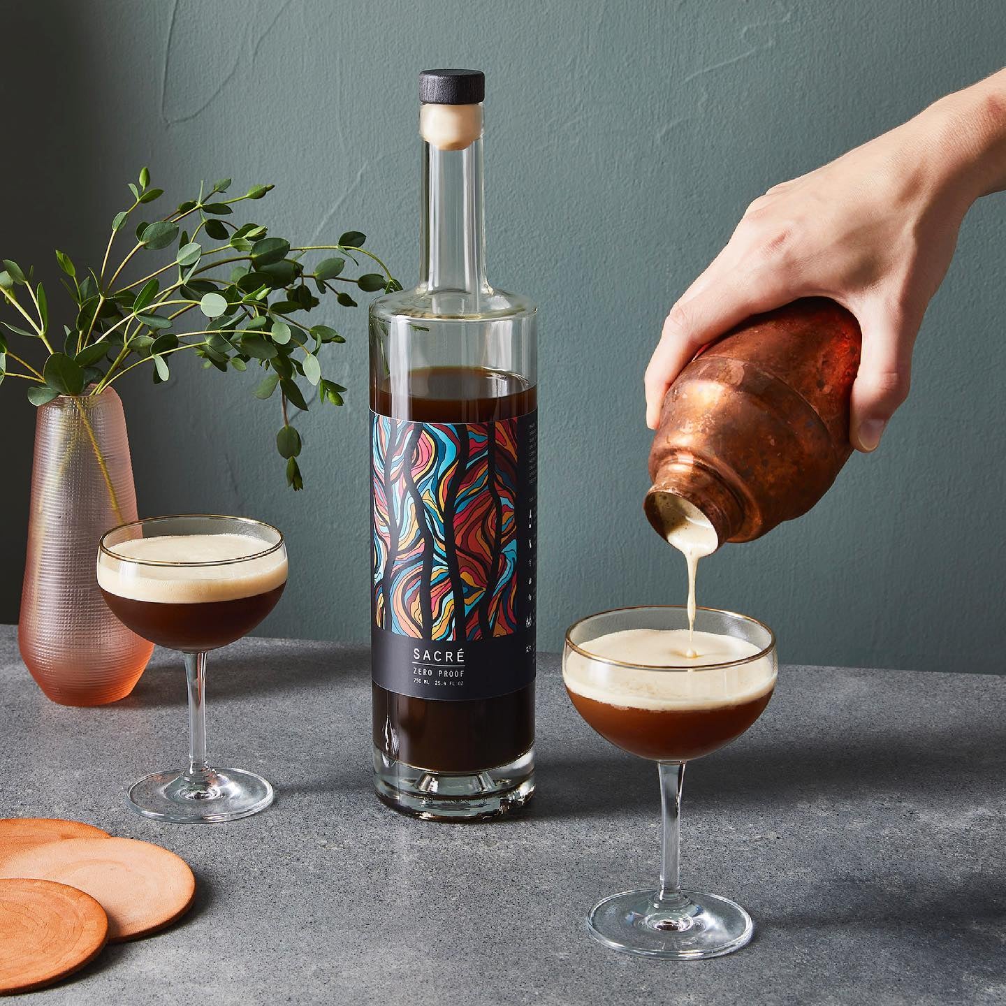 Bottle of coffee liqueur with a colorful label, being poured into a glass by a hand, with a plant and coasters in the background.