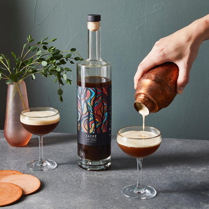 Bottle of coffee liqueur with a colorful label, being poured into a glass by a hand, with a plant and coasters in the background.