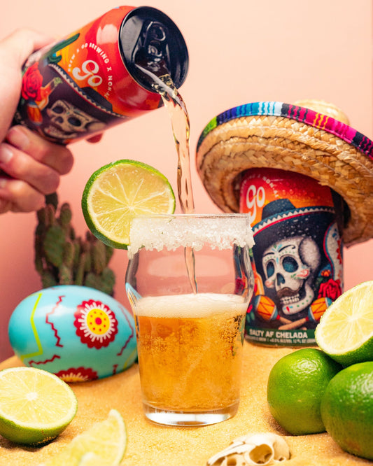 Person pouring beer from a can into a glass with limes and a colorful sombrero in the background.
