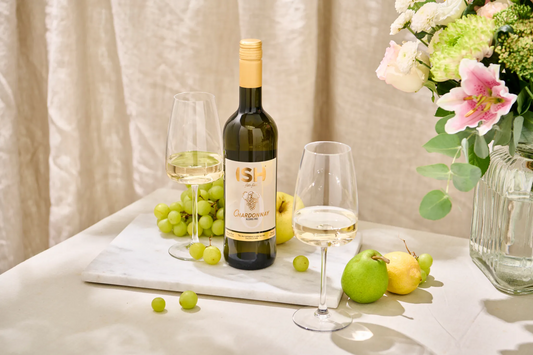 Bottle of wine with two glasses and fruits on a table with a floral arrangement.