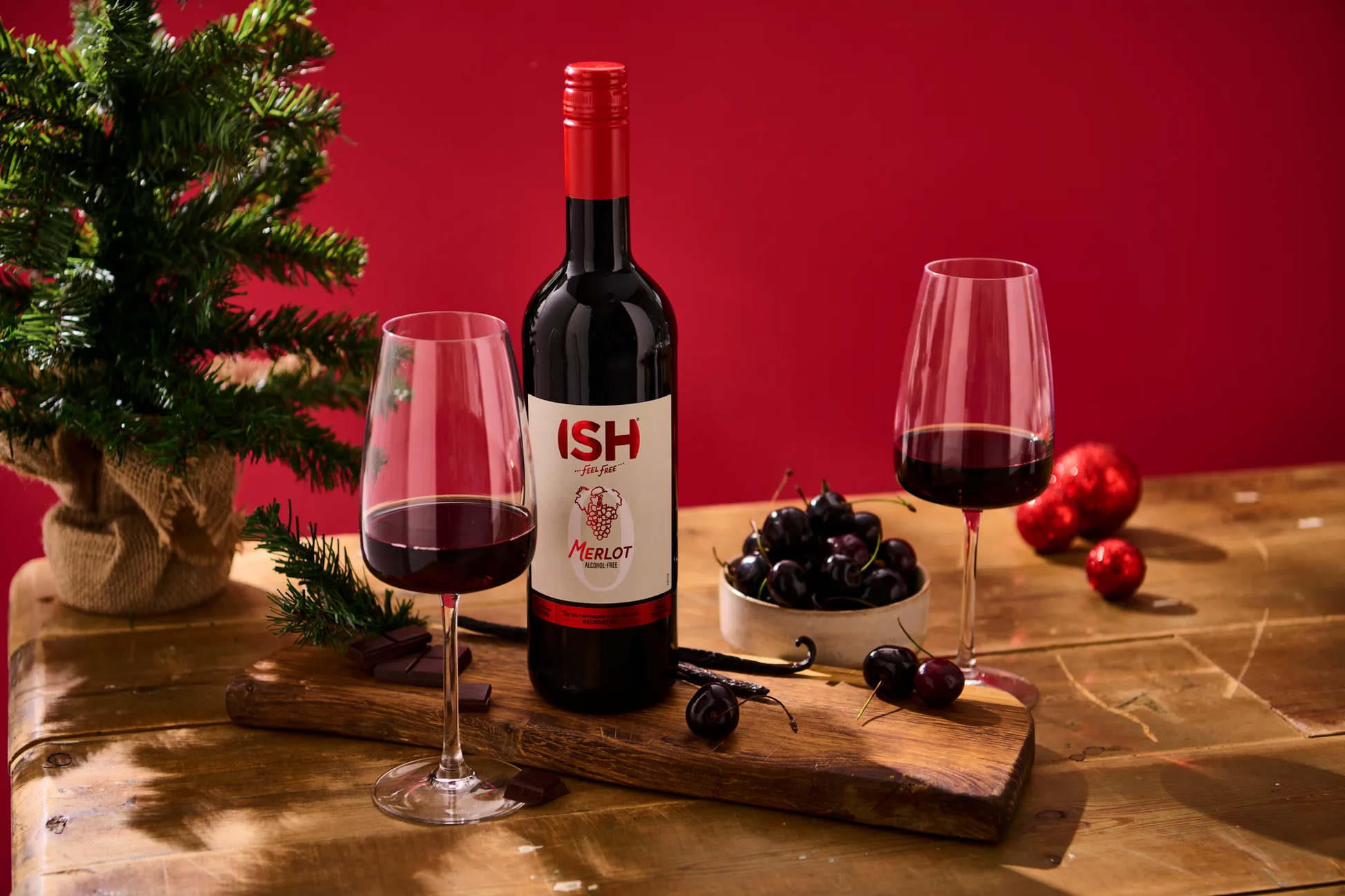 Bottle of red wine with 'ISH' label on a wooden board with two glasses, grapes, and a small Christmas tree against a red background.