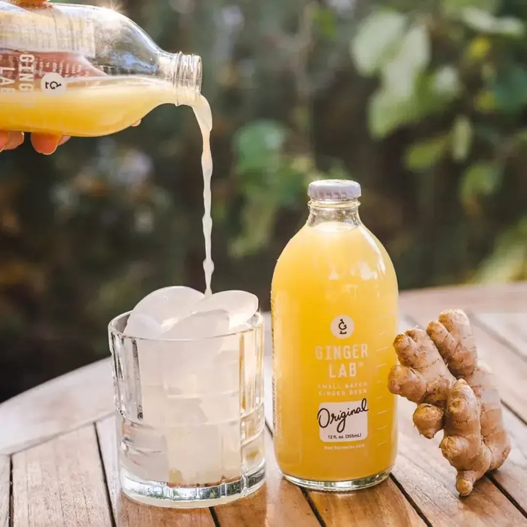 Person pouring ginger juice into a glass with ice on a wooden table, with a bottle of Ginger Lab juice and a piece of ginger in the background.