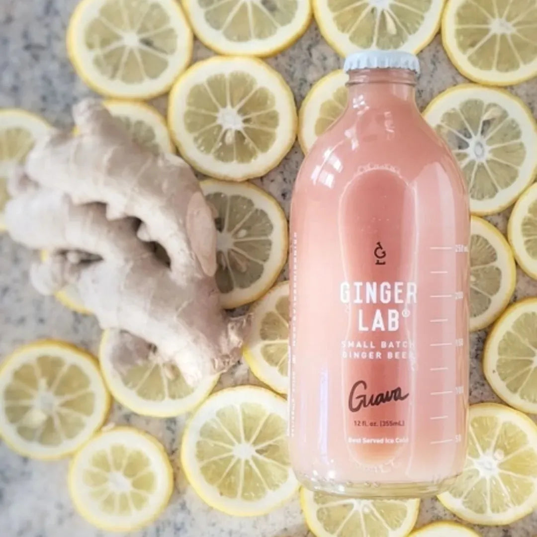 Bottle of Ginger Lab drink surrounded by sliced lemons and a piece of ginger on a gray surface.