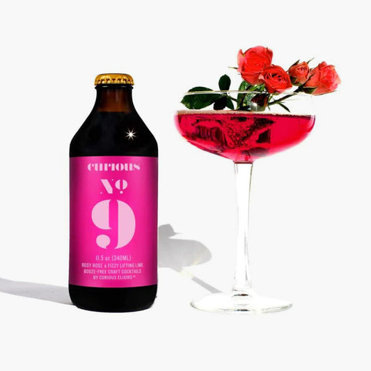 Bottle of 'curious No 9' with a pink label next to a cocktail glass filled with a red drink and flowers.