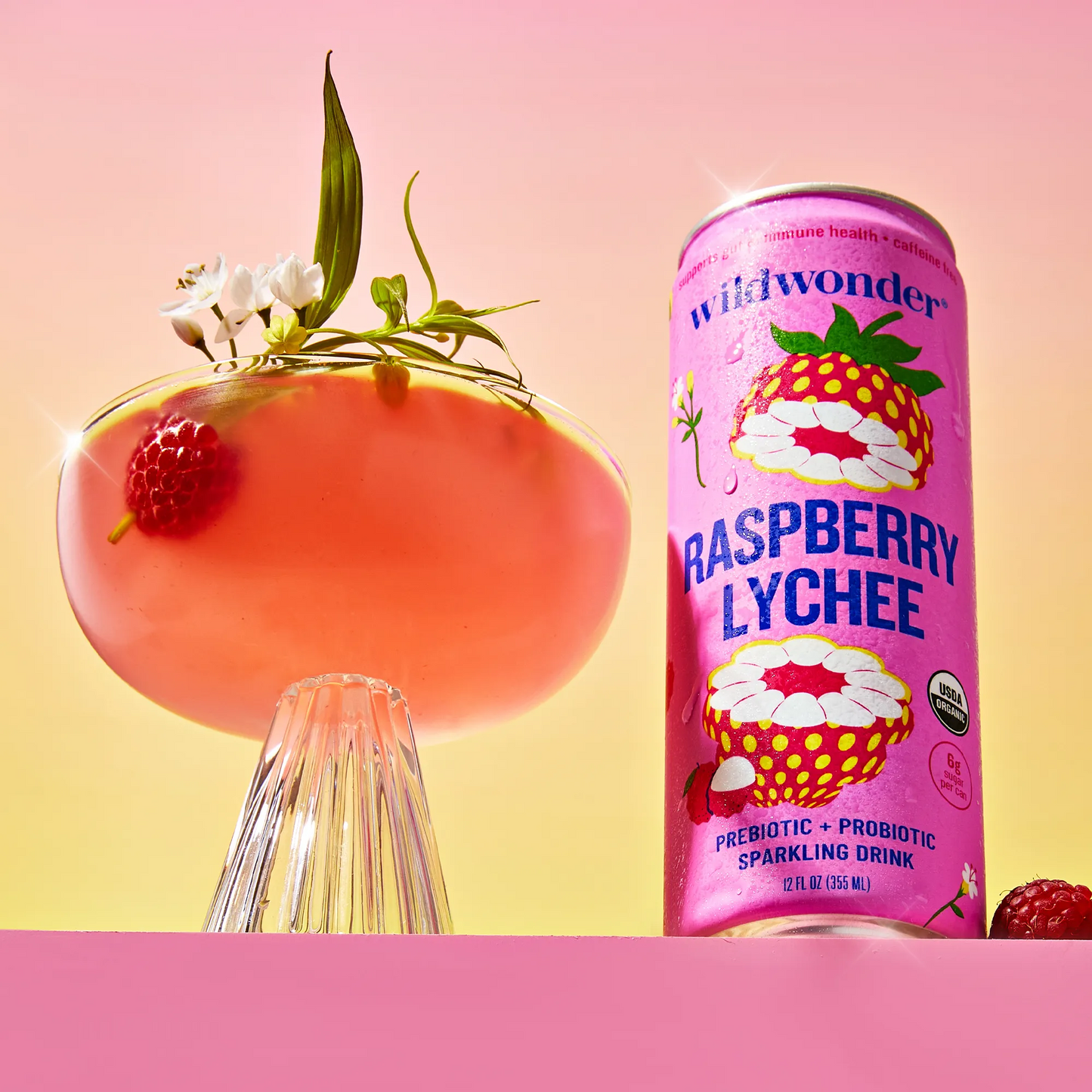 Pink can of Wildwonder Raspberry Lychee sparkling drink with a cocktail garnished with raspberries and flowers on a gradient background.