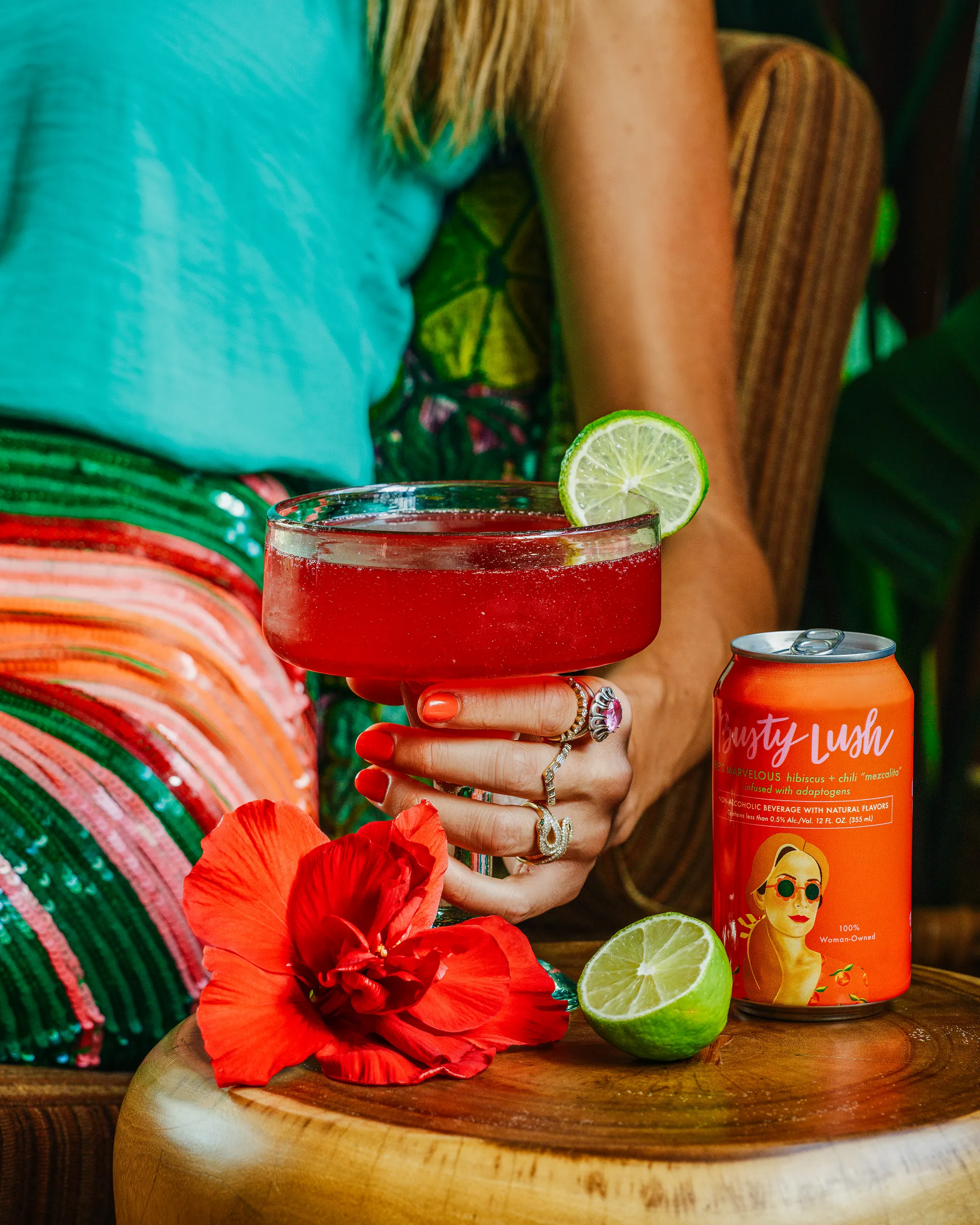 Person holding a cocktail with a can of 'Busty Lush' drink on a table with a colorful background