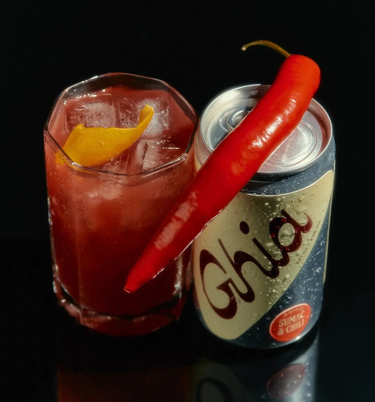 Red cocktail with a red chili pepper garnish next to a can of Ghia on a black background