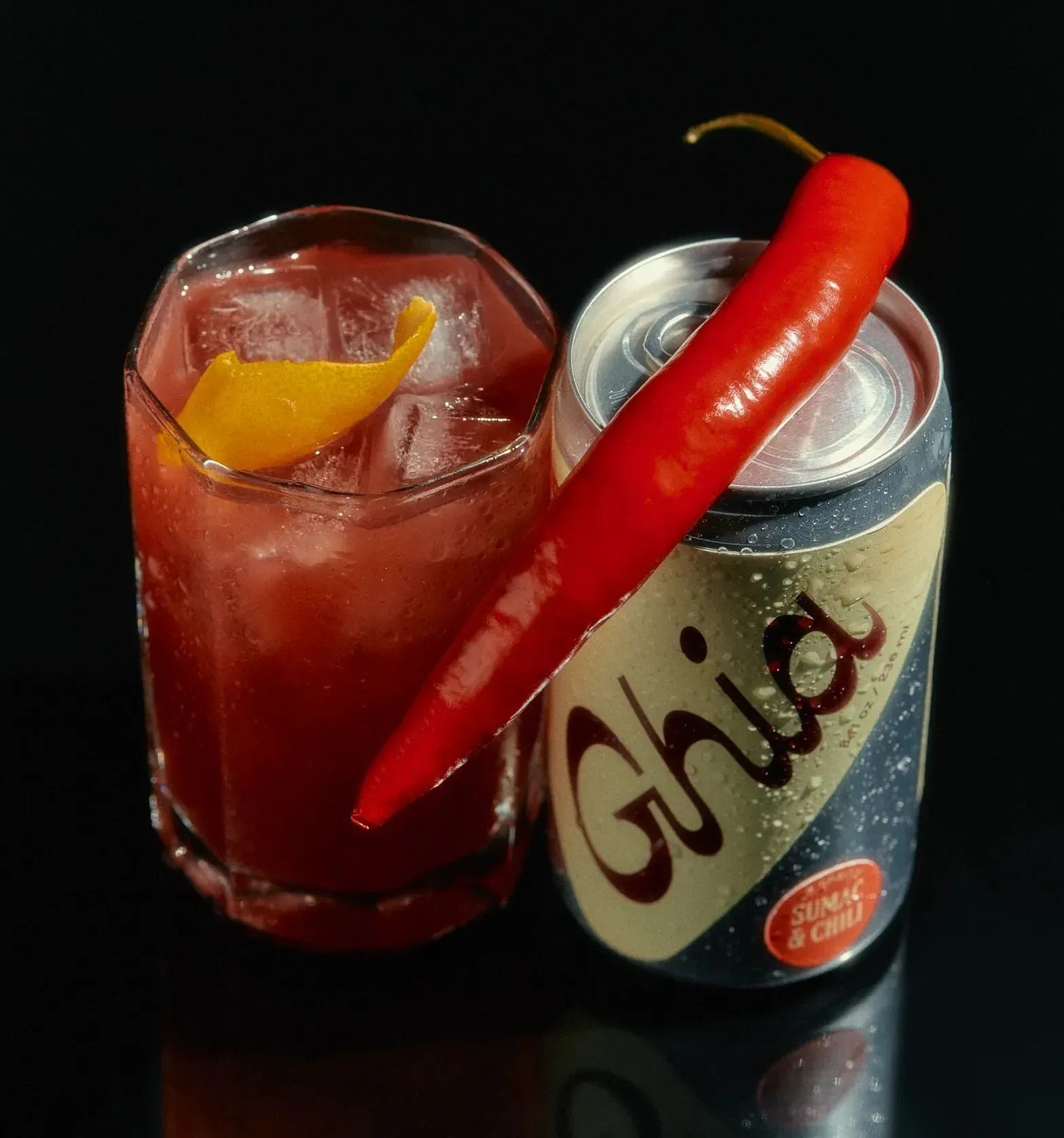 Red cocktail with a red chili pepper garnish next to a can of Ghia on a black background