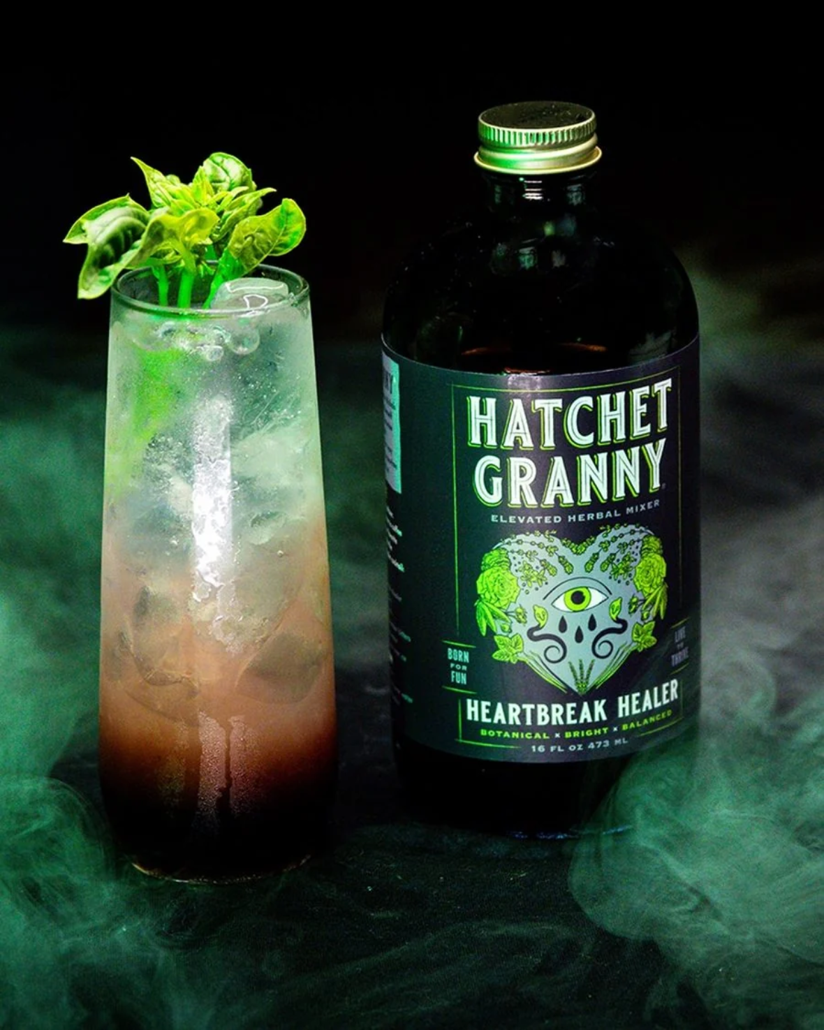 A dark bottle labeled Hatchet Granny Heartbreak Healer stands beside a tall glass filled with ice and a layered drink, garnished with green leaves, against a smoky black background.