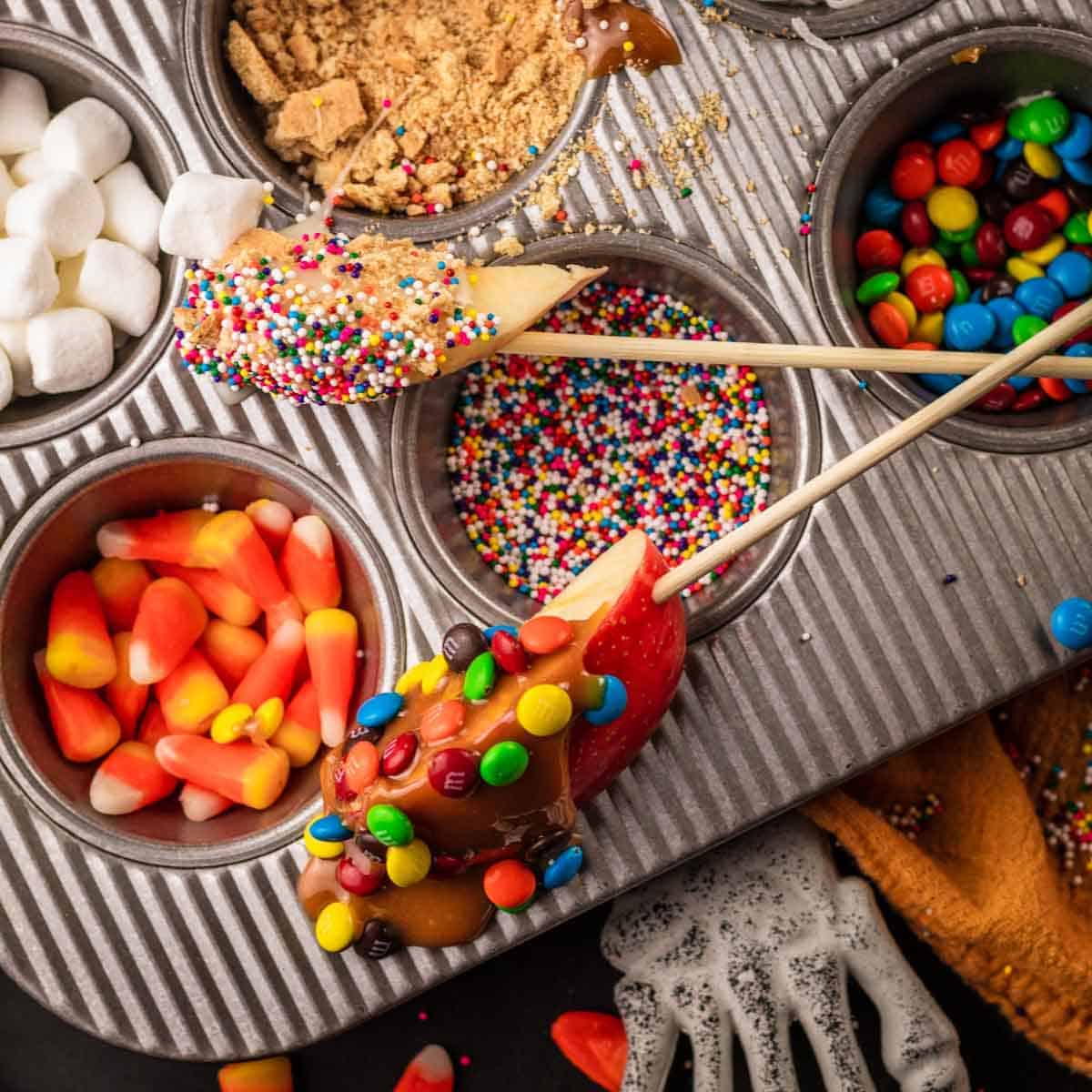 A Halloween-themed candy apple dessert being dipped in caramel and sprinkles inside a metal baking pan filled with treats, marshmallows, candy corn, M&M's, and a skeleton hand decoration.