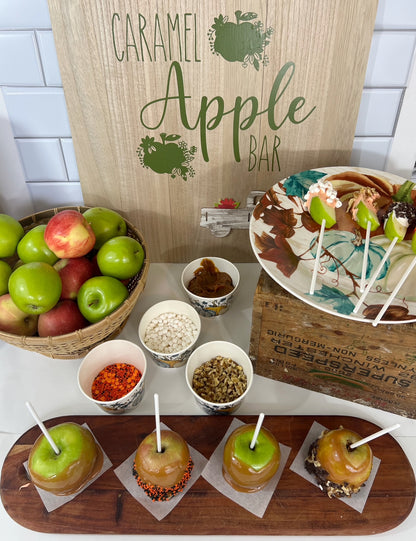 A styled caramel apple bar setup with green and red apples, bowls of toppings, lollipops, and four caramel-coated apples on a wooden board, in front of a wooden sign reading 'Caramel Apple Bar'.