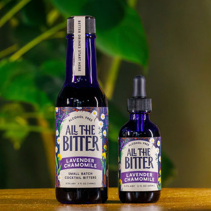 Two bottles of 'All The Bitter' lavender chamomile bitters on a wooden surface with a blurred green background.
