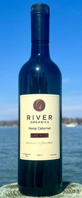 A bottle of River Organics Hemp Cabernet red wine stands on a stone surface with a blurred outdoor background of blue sky and water.