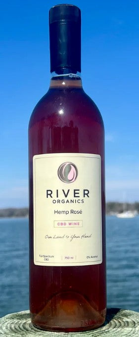 A bottle of River Organics Hemp Rosé wine stands outdoors on a stone surface with water and a blue sky in the background. The bottle is dark glass with a cream label and a black cap.