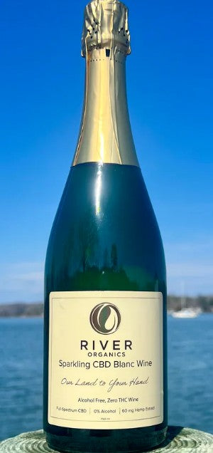 A bottle of River Organics Sparkling CBD Blanc Wine, featuring a dark green glass bottle with a gold foil neck and a cream-colored label, set against a bright blue sky and water background.