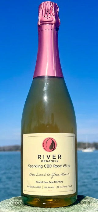 A bottle of River Organics Sparkling CBD Rosé Wine with a pink foil top and cream label, set outdoors against a blue sky and water background.