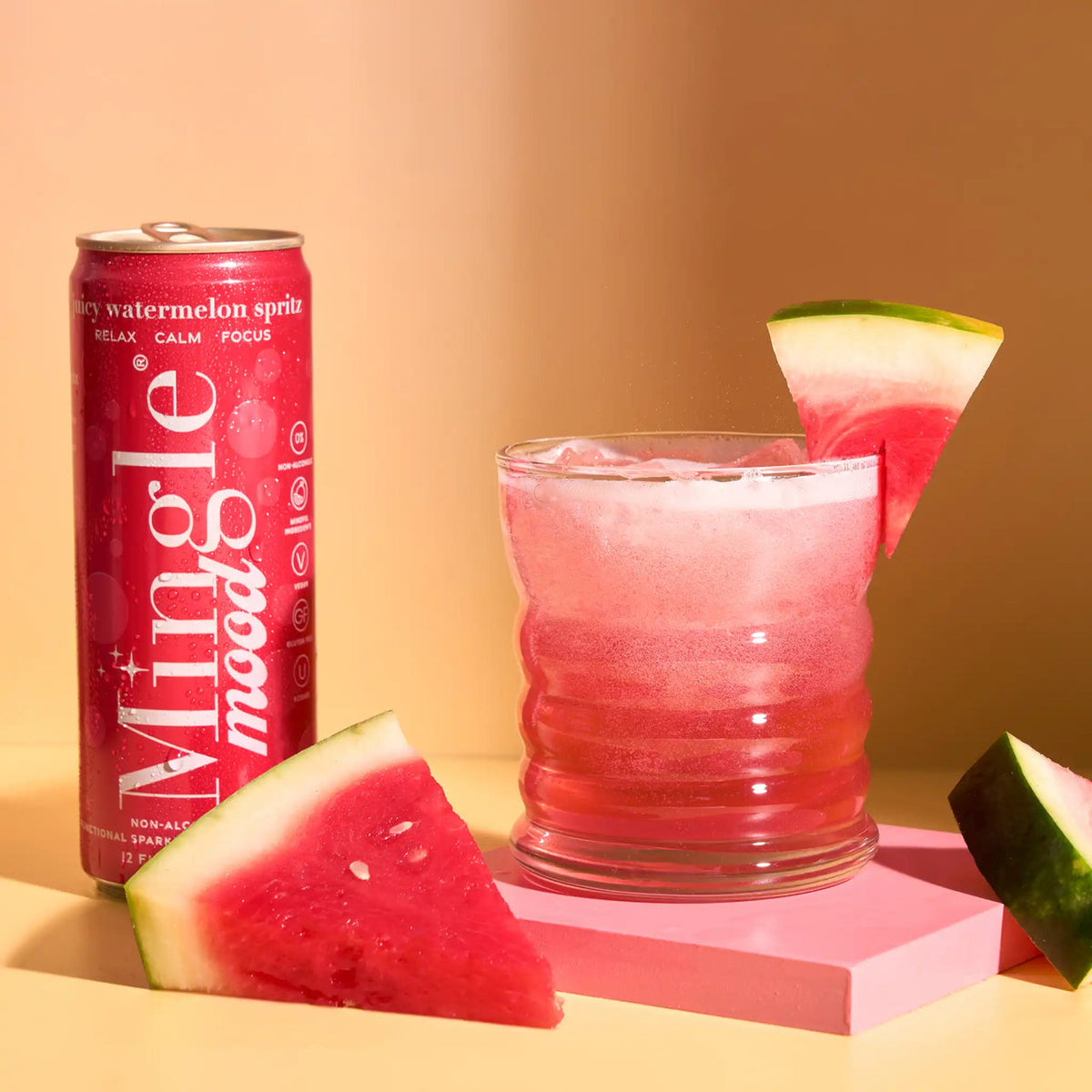 A can of Mingle Mood watermelon sprite stands next to a glass of pink sparkling drink garnished with a watermelon slice, with additional watermelon wedges on a warm beige surface against a peach background.